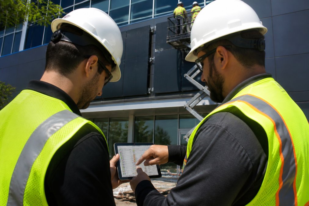 Two installation crew members in high-visibility vests reviewing a tablet displaying a scheduling app at the base of a commercial building exterior where large signage panels are being mounted, midday natural light