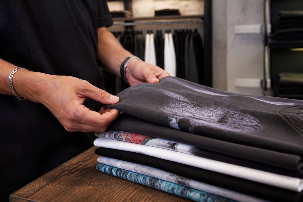Close-up of a man's hands feeling the thick cotton fabric of a folded stack of premium graphic t-shirts in a modern, minimal clothing store