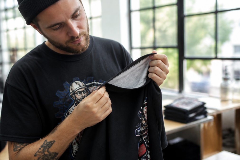 A person examining the fabric and stitching of a bold graphic t-shirt under natural daylight near a window, turning it inside out to inspect the seams