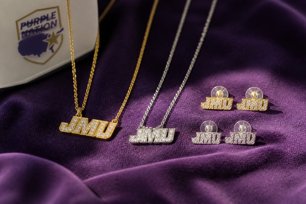 Close-up of JMU necklaces and earrings displayed on soft purple fabric, showing engraved block JMU logos and delicate gold and silver finishes catching natural light