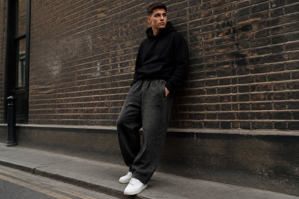 Street style photograph of man wearing oversized grey sweatpants with fitted black hoodie tucked partially into the waistband, white trainers, standing against a brick wall with urban background