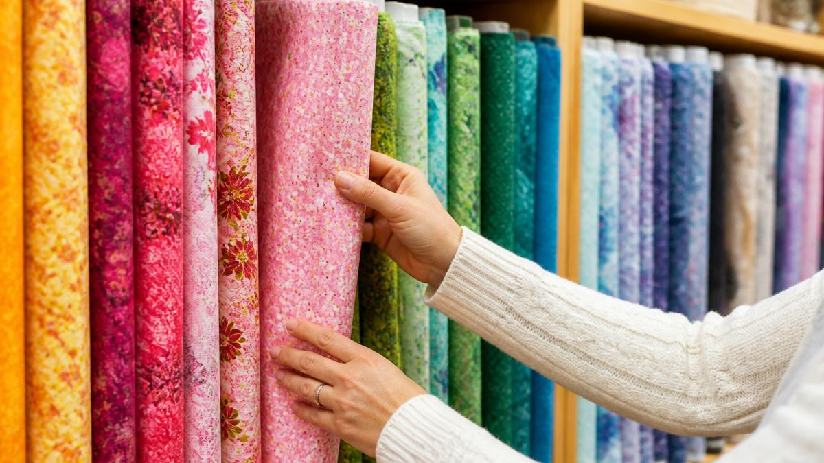 Close-up of hands touching and examining colorful quilting fabric bolts arranged on wooden shelves, with warm natural light highlighting the texture and patterns of the cotton materials