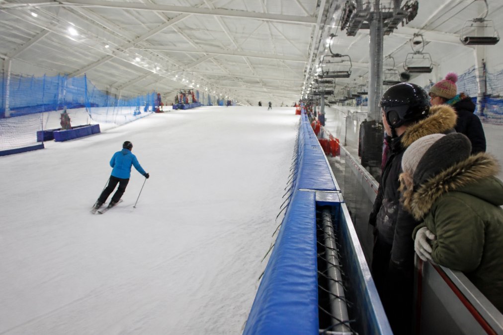 Skier navigating down an indoor snow slope with spectators watching from the side, showing real snow and the indoor facility environment with bright lighting