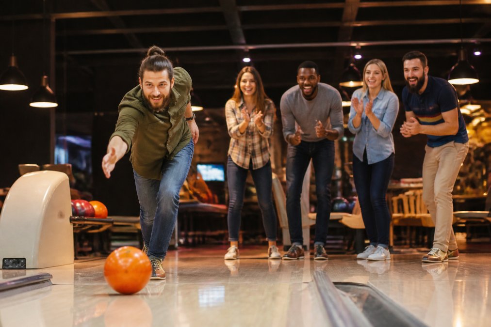 Group of adults enjoying ten-pin bowling, with someone mid-swing and others cheering nearby, showing the fun social atmosphere and bright venue lighting