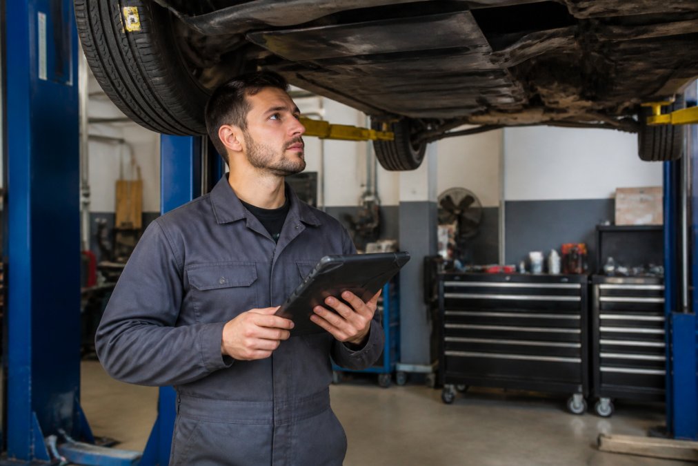 A certified automotive technician in a clean uniform inspecting a vehicle on a lift in a well-lit independent repair shop, diagnostic tablet in hand, with tool chests visible in the background