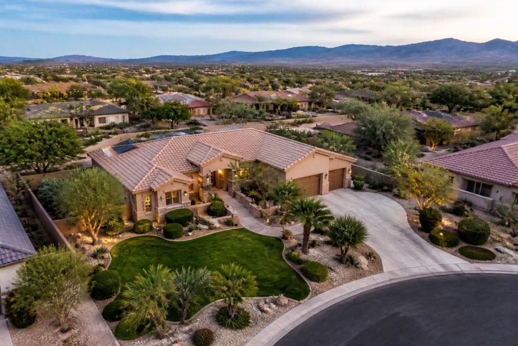 Aerial view of a residential property with manicured landscaping, visible driveway, and surrounding neighborhood visible from above, showing professional drone photography perspective