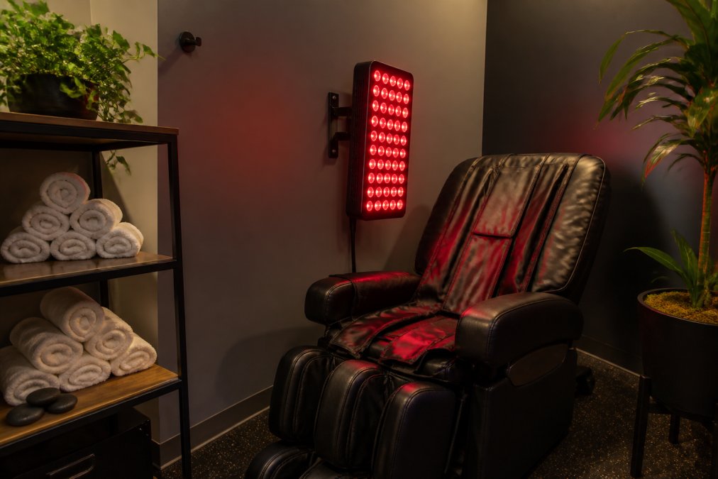A compact, well-organized recovery corner inside a small gym, featuring a red light therapy panel mounted on the wall beside a compression chair, with a clean towel rack and dimmable ambient lighting