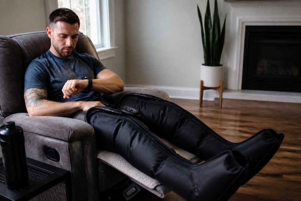 Athlete in recovery boots at home with a biometric tracking device visible on their wrist, demonstrating integrated recovery routine in a comfortable home setting
