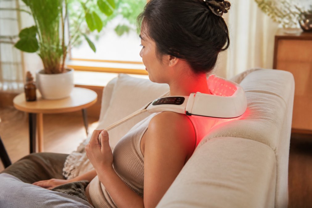 Person using red light therapy device on neck and shoulders while sitting comfortably at home with warm natural lighting from window
