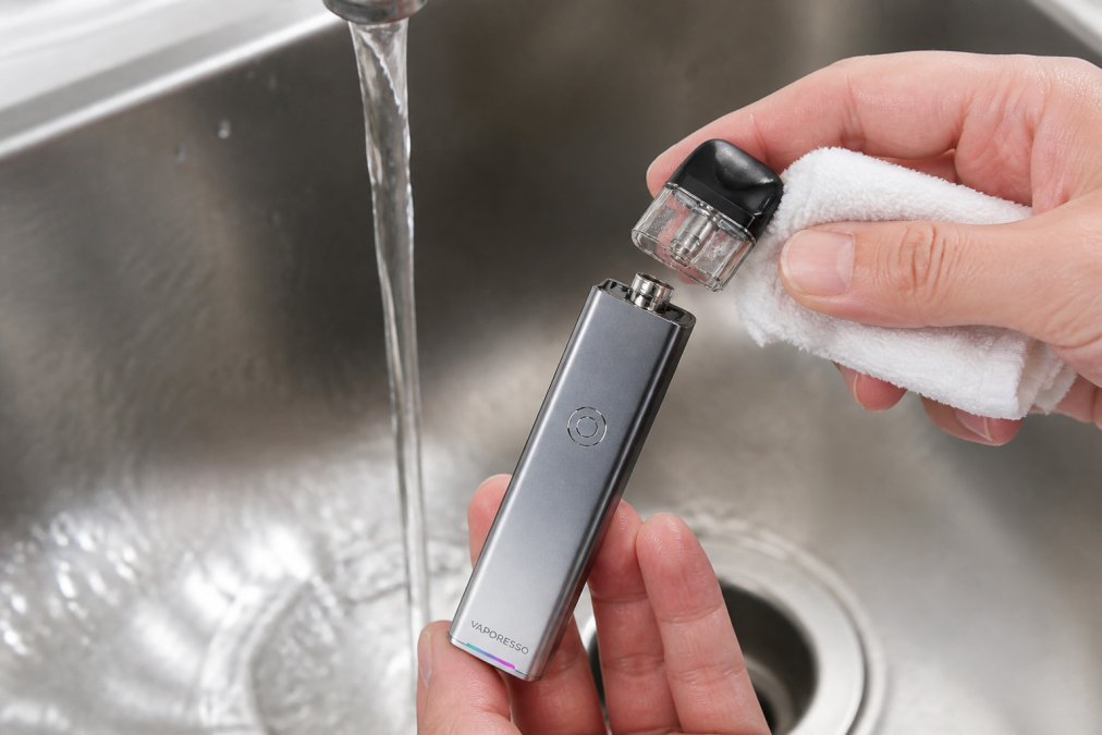 Hands carefully cleaning a vape pod over a sink with soft cloth and warm water, showing the top-fill system and coil detail