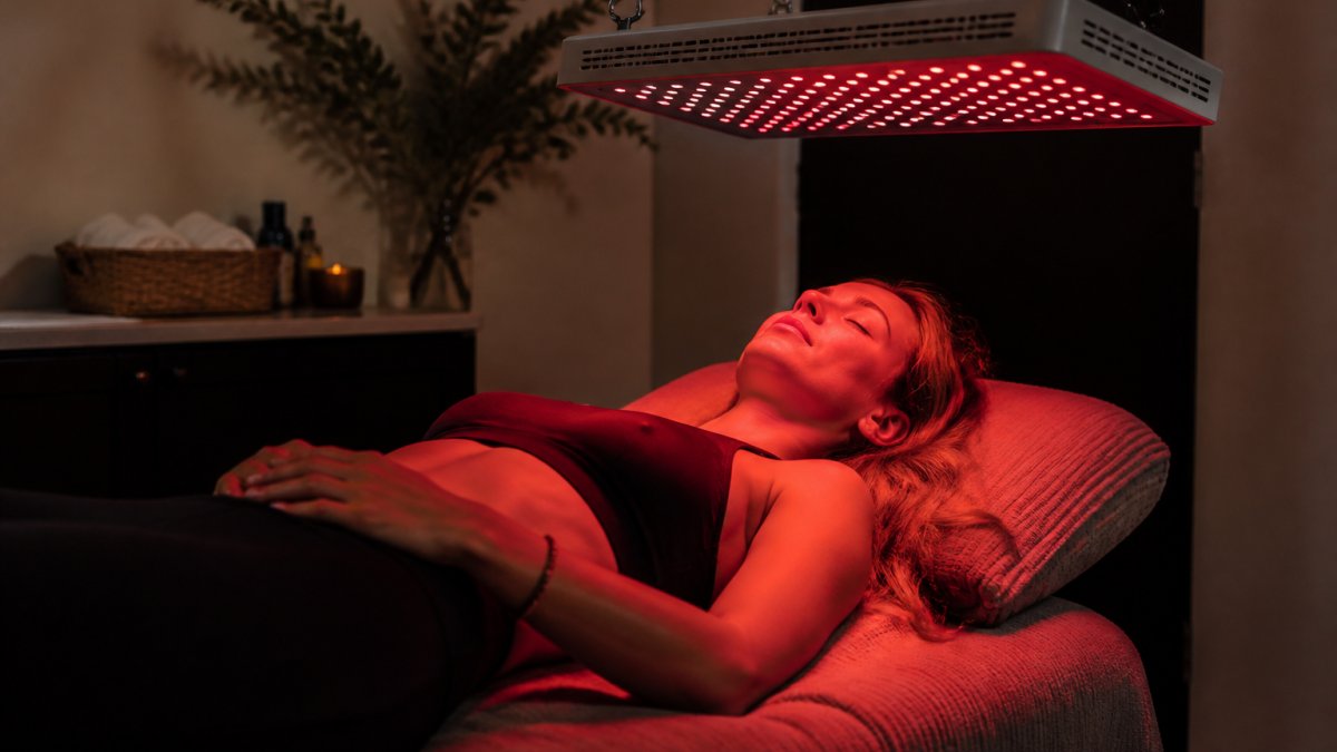 A woman resting comfortably on a treatment table under a red light therapy panel in a calm, dimly lit wellness studio