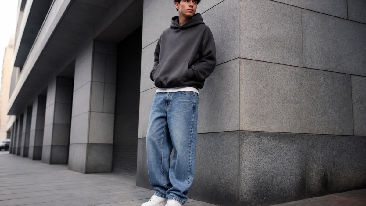 Urban male model wearing relaxed fit jeans with oversized hoodie, white sneakers, and minimal accessories standing against modern city street architecture with natural daylight