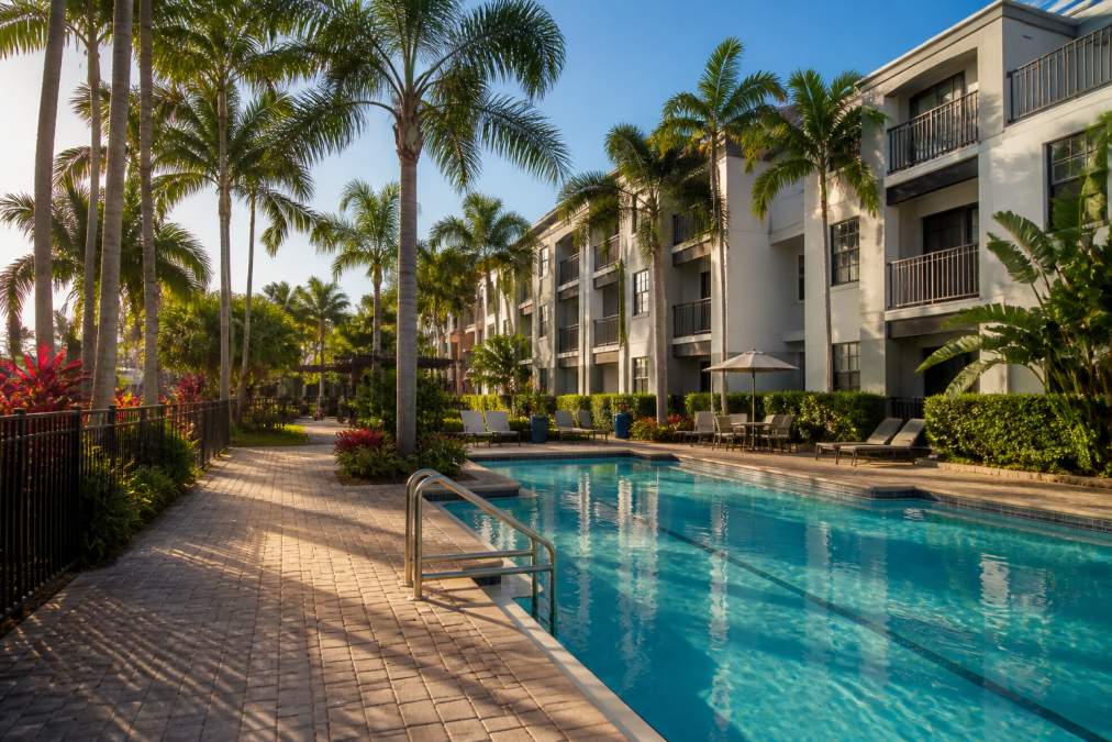 A modern garden-style apartment building in Boca Raton, Florida with lush tropical landscaping, a resort-style pool in the foreground, and palm trees lining the walkway in warm afternoon light