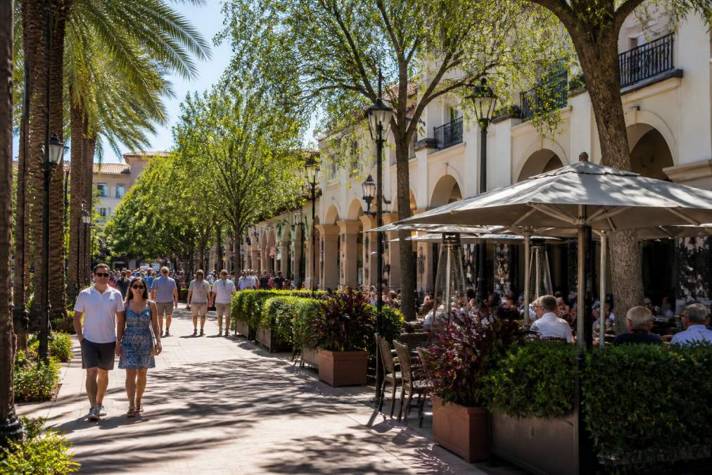 Vibrant street scene at Mizner Park with outdoor dining patios, pedestrians strolling along tree-lined plaza, upscale shops and restaurants visible in background on a sunny afternoon