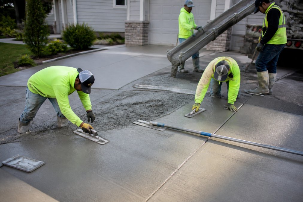 Construction crew carefully pouring and finishing fresh concrete on a residential driveway with professional trowels and equipment visible under bright daylight