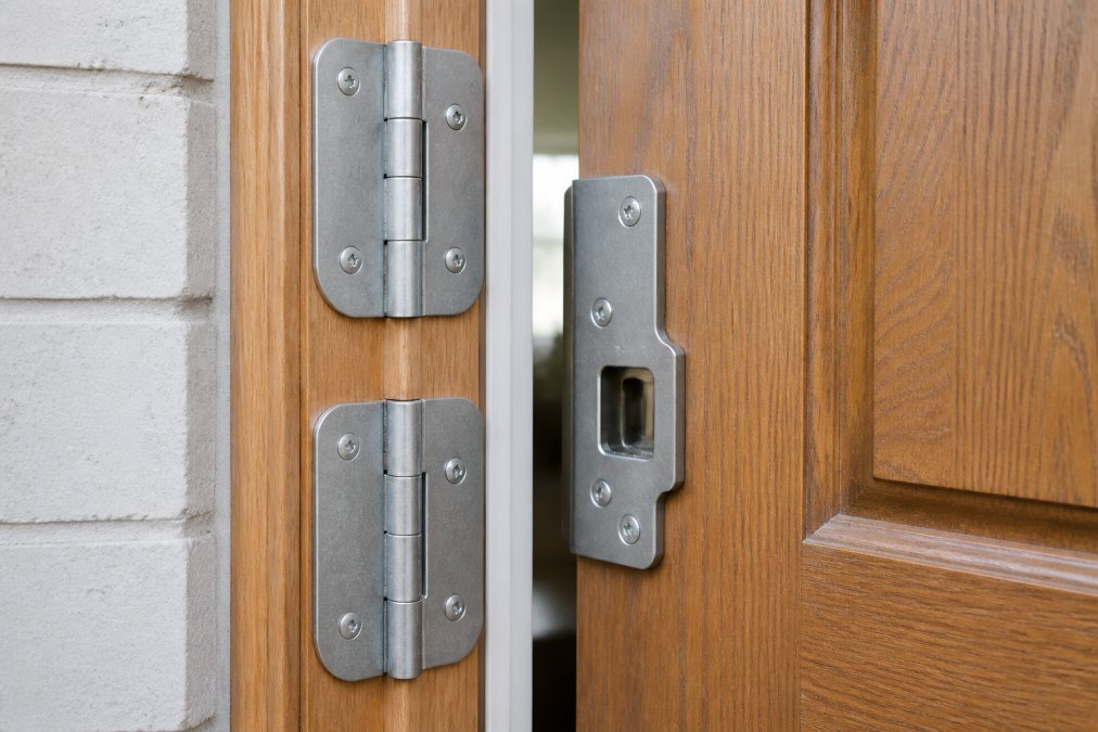 Close-up of galvanized steel door reinforcement hardware showing hinge shields and strike plate reinforcement installed on wooden door frame with natural daylight