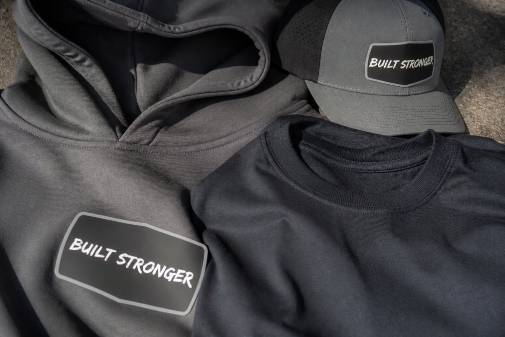 Close-up flat lay of EVRMORE Built Stronger heavyweight hoodie and tee showing fabric texture, bold graphic details, reinforced seams, and construction quality in natural light