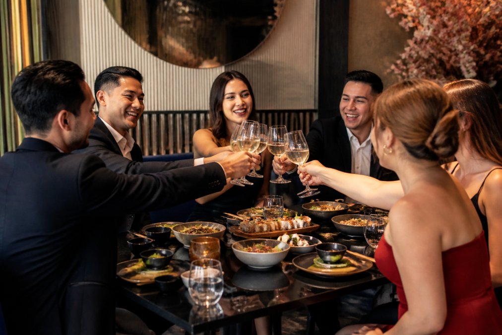 A group of elegantly dressed guests enjoying a festive dinner together at Pangaea Grill, sharing plates of Asian fusion cuisine with warm ambient lighting, raising glasses in a toast while laughing