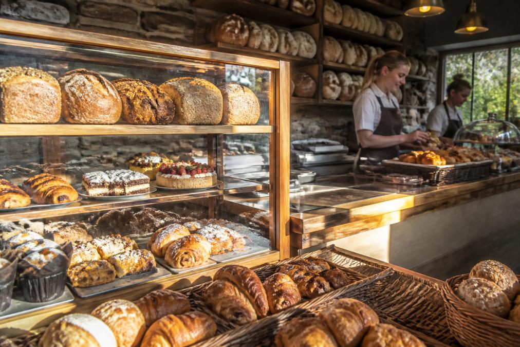 Warm interior of a traditional Scottish bakery displaying fresh baked goods in window cases, with natural morning light streaming across shelves of bread, pastries, and cakes, and a welcoming counter with staff