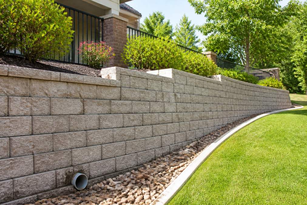 A wide shot of a professionally built segmental concrete block retaining wall on a residential slope, with clean landscaping and a visible drainage pipe outlet at the base
