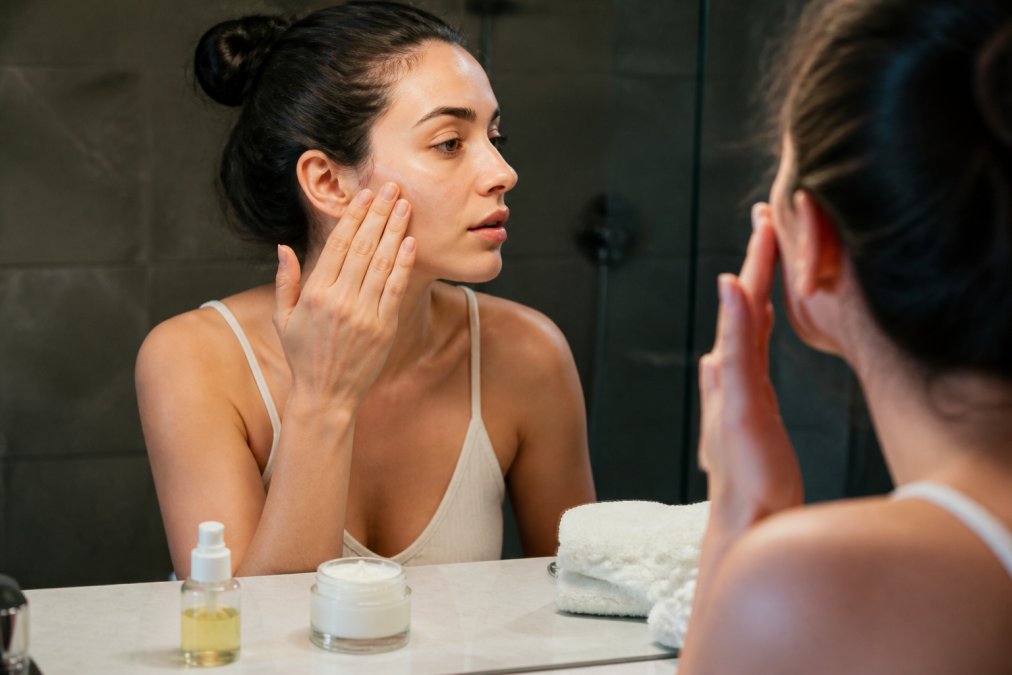 Close-up of a woman's hands applying moisturizer to her face with a small pump bottle of serum and a jar of cream on a bathroom counter