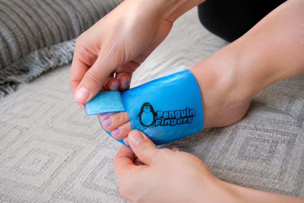 Hands adjusting the compression straps of a cold wrap around a person's foot, demonstrating proper tightening technique with the wrap conforming to the foot's contours, showing the adjustable velcro straps being secured