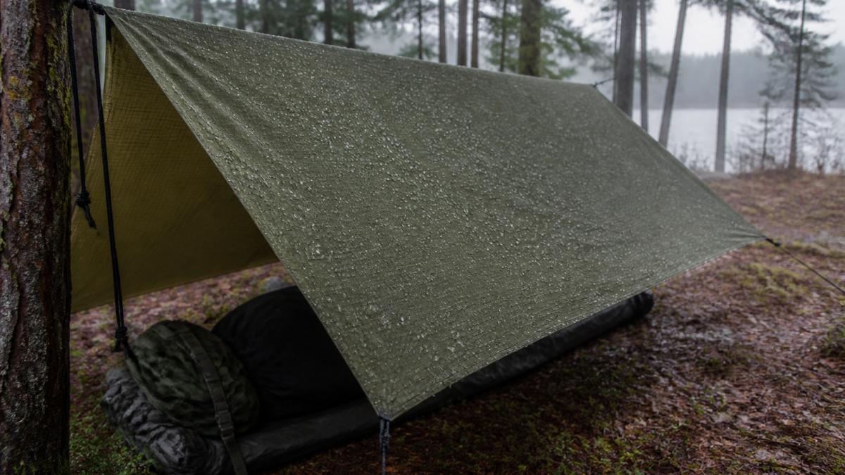 A ripstop tarp deployed in a rugged outdoor camping setting with visible reinforced weave pattern and tight grid structure under natural daylight, showing moisture beading on the surface