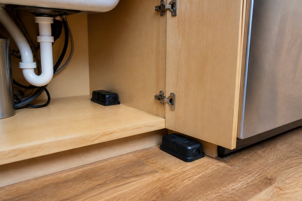 Close-up view of kitchen corner showing roach bait stations positioned in cracks and crevices near cabinet hinges, under sink pipes, and along appliance edges with natural kitchen lighting