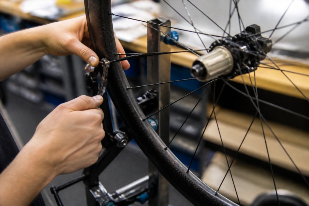 Skilled technician's hands carefully truing a carbon wheel spoke by spoke, adjusting tension with precision tools in a well-lit workshop, showing close-up detail of the hub and spoke interface