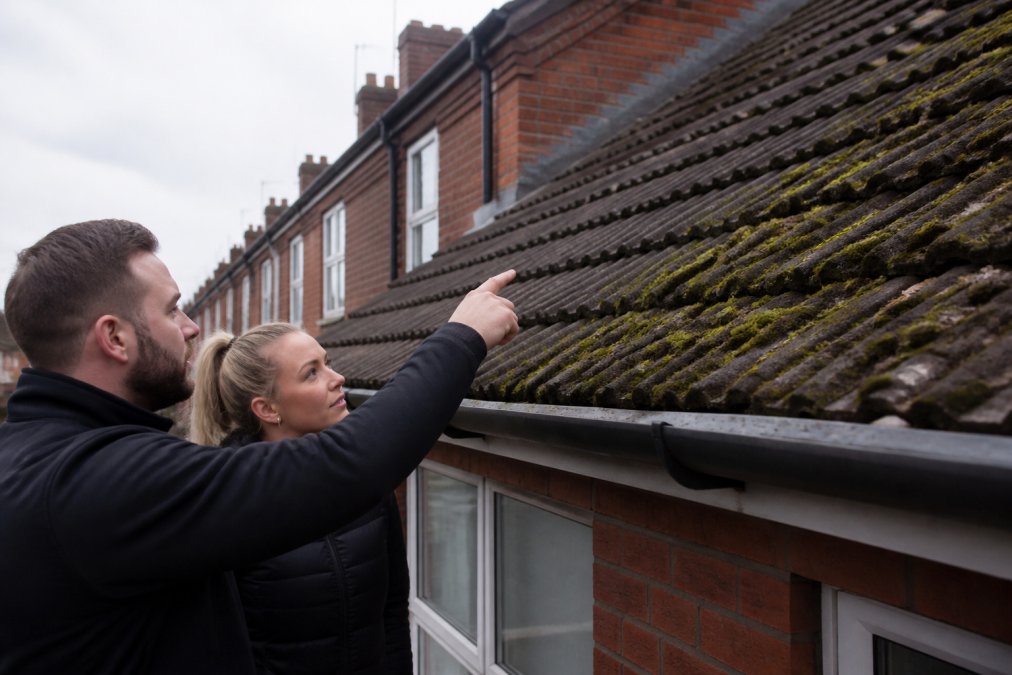 A terraced house roofline in Bolton with thick green moss along the north-facing tiles and a clear gutter below, overcast daylight
