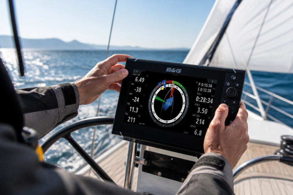Professional sailor's hands adjusting a Garmin chartplotter display mounted at the helm during daylight sailing, with ocean horizon and white sails visible in the background