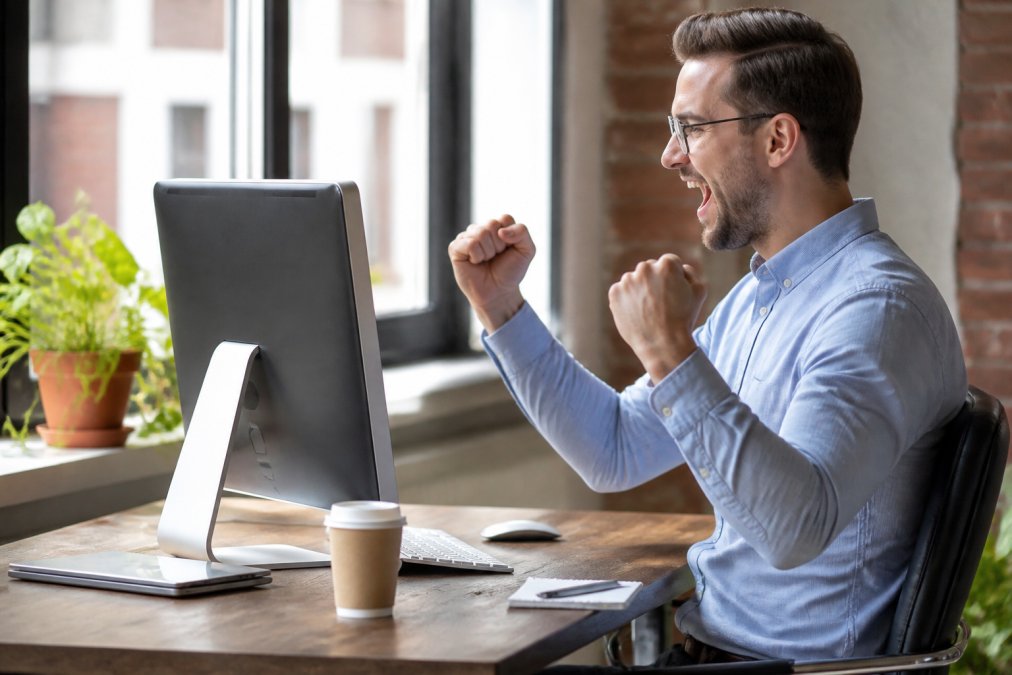 Remote sales professional celebrating achievement while viewing real-time leaderboard on desktop monitor, coffee cup nearby, natural office lighting from window