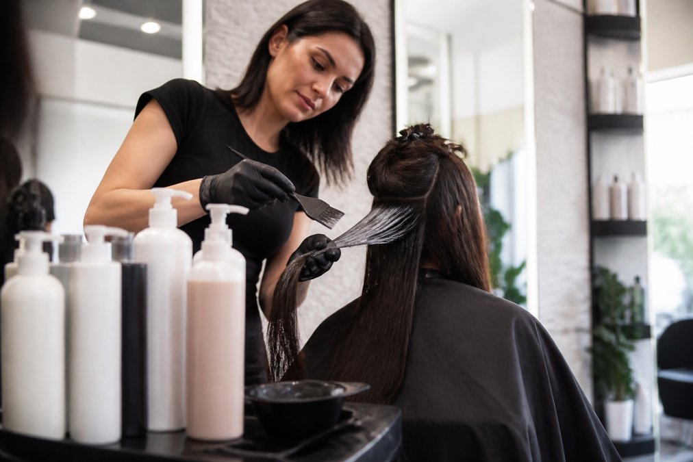 A professional hairstylist applying a keratin treatment to a client's hair in a bright, modern salon, with product bottles visible in the foreground