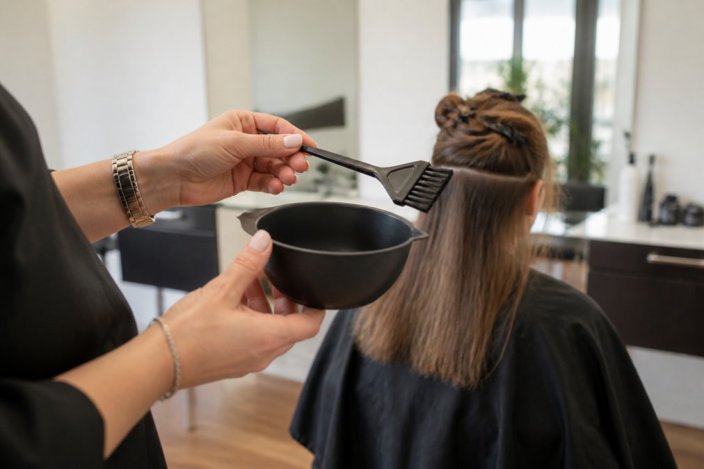 A professional colorist's hands holding a stain-resistant color bowl and a feathered tint brush, with a client's hair sectioned in the background of a bright, modern salon studio