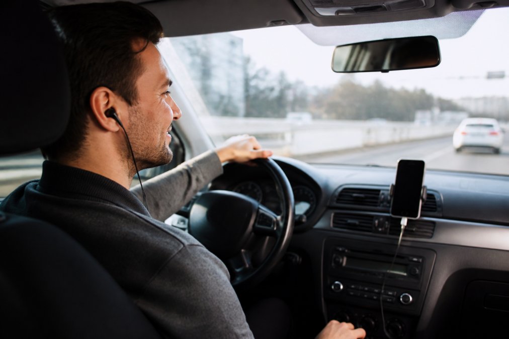 A driver in a car with a smartphone mounted on the dashboard, earbuds in, smiling as they listen to an audiobook during a commute