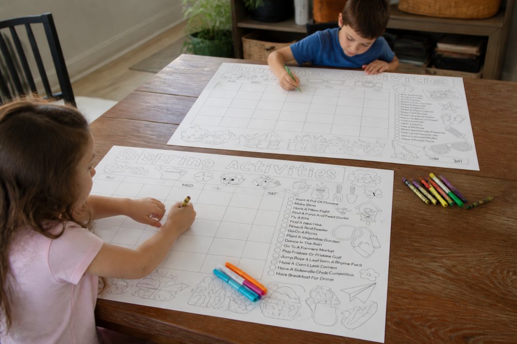 Young child sitting at table coloring on a large oversized coloring sheet with markers and crayons, older sibling engaged in their own section of the same sheet nearby
