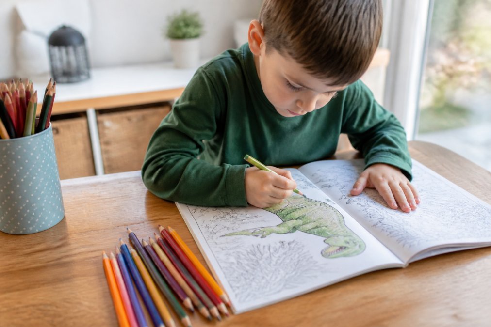 A young child sitting at a wooden table, deeply focused on coloring a detailed dinosaur page in an activity book, with colored pencils scattered nearby in a bright, naturally lit room