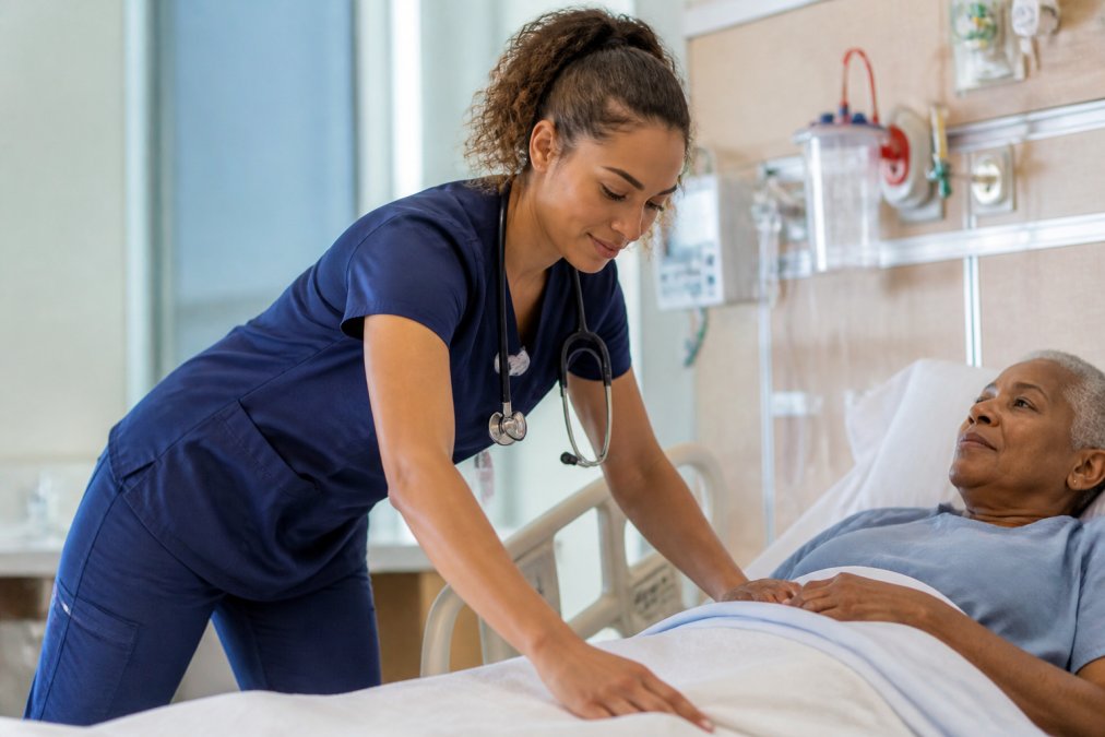Nursing student wearing modern-fit scrubs during a clinical rotation, bending and reaching to assist a patient, demonstrating mobility and comfort during active movement in a hospital setting