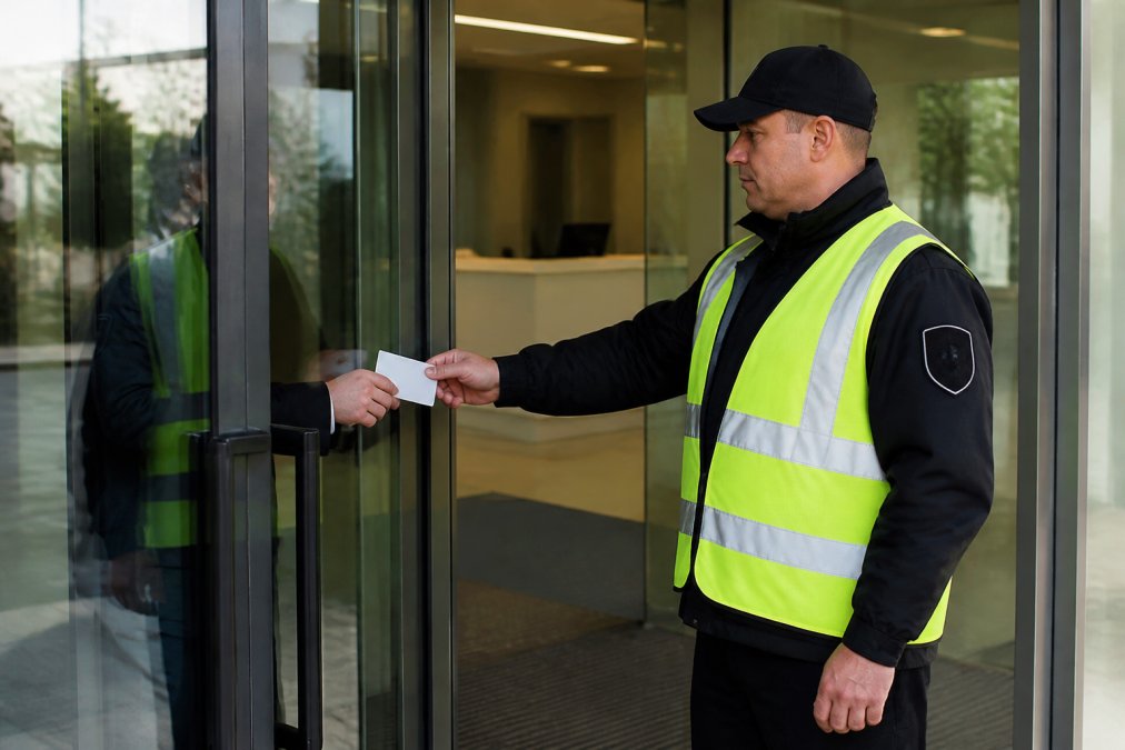 Professional security guard in formal dark uniform and high-visibility safety vest conducting access control at commercial building entrance, checking credentials at glass doors with modern office interior visible