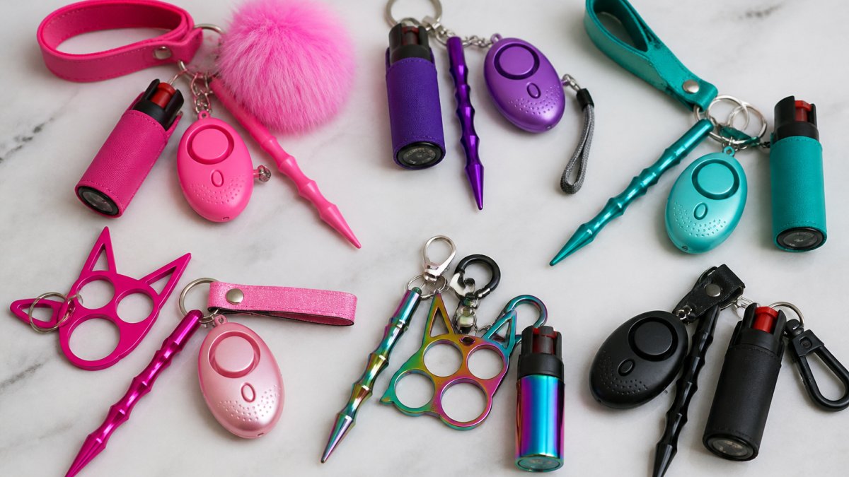 Flat-lay arrangement of multiple colorful self-defense keychain sets displayed on a neutral surface, showing various designs including hot pink, purple, teal, and rainbow options with visible components like personal alarms, pepper spray bottles, and metal tools