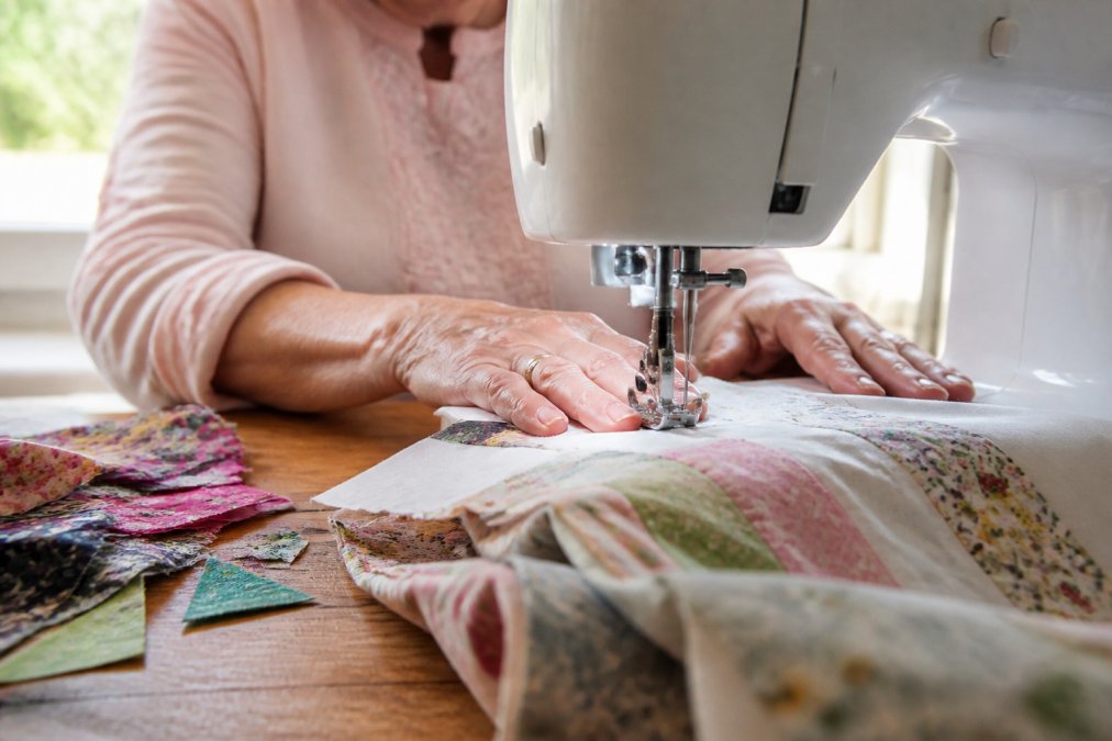 Close-up of an older woman's hands guiding fabric through a sewing machine on a wooden quilting table, with colorful fabric scraps and a walking foot attachment visible, soft natural light from a nearby window