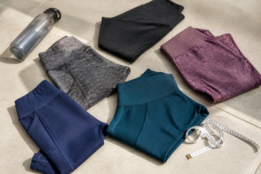 A close-up flat-lay of several folded workout leggings in different fabric textures and waistband styles arranged on a light gym floor, with a measuring tape and a water bottle positioned nearby