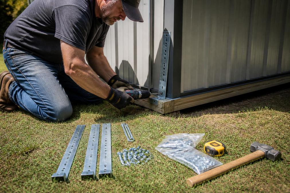 A person in work gloves kneeling beside a metal shed's base, installing a steel hurricane strap with a drill against the shed's lower frame, with anchor hardware and tools laid out on the grass in the foreground, afternoon sunlight casting shadows across the metal panels
