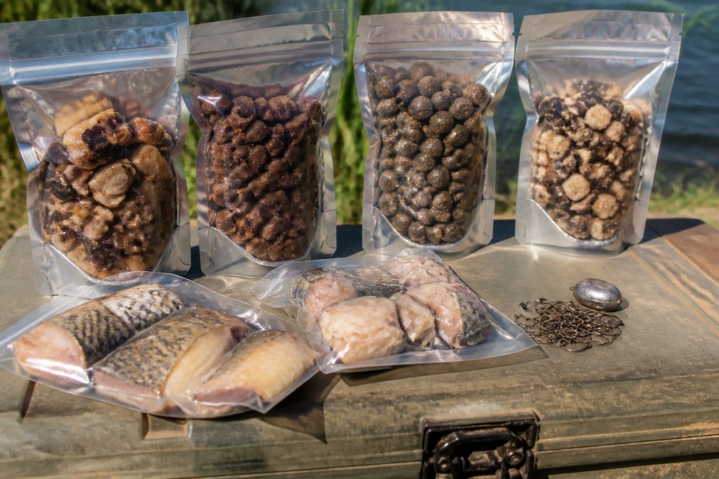 Close-up of multiple resealable bags containing preserved bait chunks and nuggets arranged on a weathered tackle box, with fishing hooks and preserved carp pieces visible in natural daylight