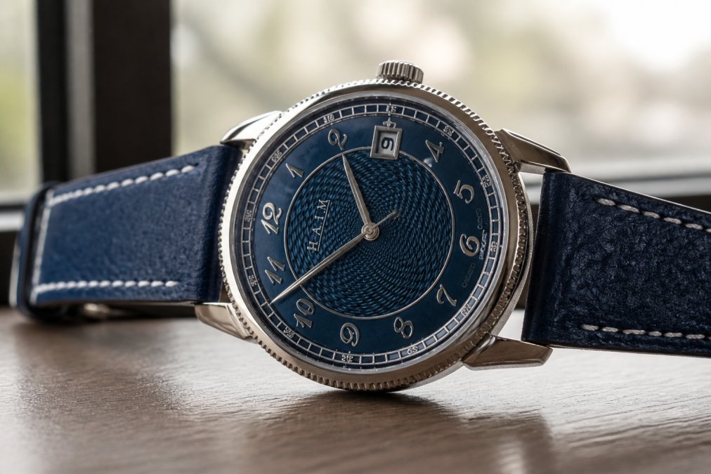 Titanium watch case with hand-finished surfaces showing brushed and polished textures, coin-edge bezel detail, and ornate dial decoration visible through crystal under natural window lighting