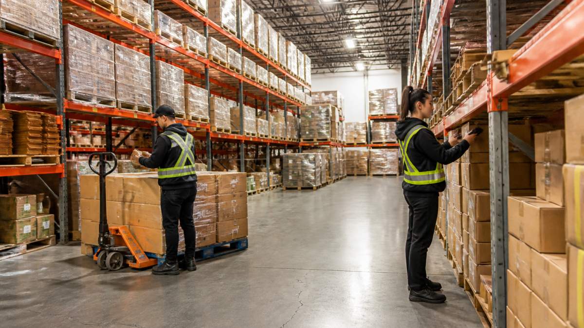 Warehouse workers organizing and scanning inventory boxes on shelves in a modern fulfillment center with bright overhead lighting and organized pallet storage