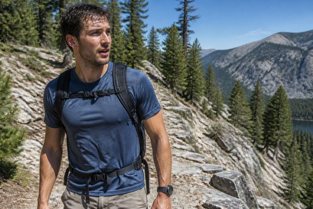 A male hiker wearing a lightweight technical shirt on a rocky mountain trail, visibly sweating but looking comfortable, surrounded by rugged terrain and pine trees in bright midday sunlight