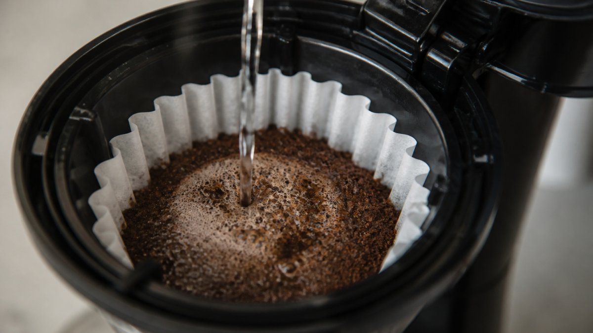 Close-up of freshly ground single origin coffee beans in a filter basket of an automatic drip machine, with hot water being poured, showing the bloom phase where water first contacts the grounds