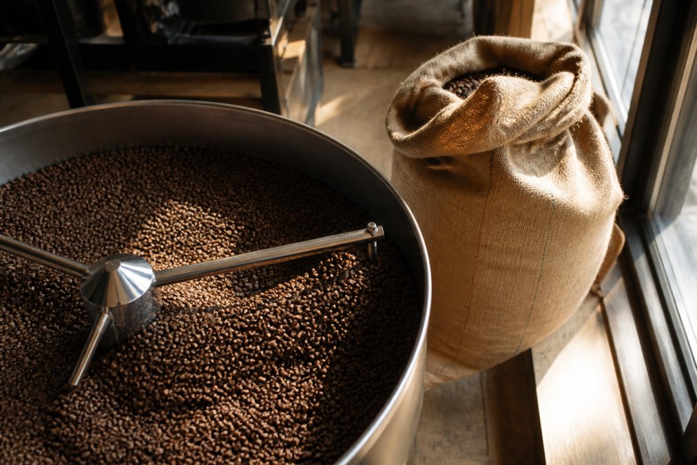 Freshly roasted single origin coffee beans cooling in a metal tray beside a burlap sack in a sunlit roastery