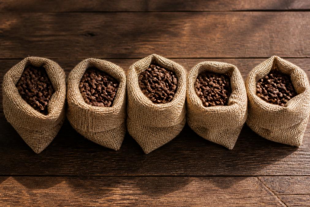 Flat lay of five small burlap bags of whole coffee beans arranged on a rustic wooden surface, each labeled with a different country of origin tag, warm natural side lighting casting soft shadows